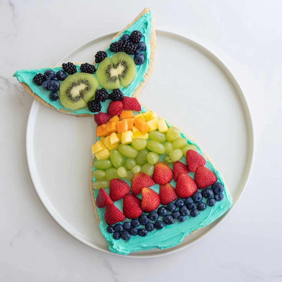 A creamy frosting base holds colorful berries and kiwi in this festive Mermaid Tail Fruit Pizza creation.