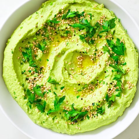 Creamy Edamame Hummus in a white bowl, drizzled with olive oil and topped with toasted sesame seeds and fresh parsley, served with pita chips and crisp veggies.  