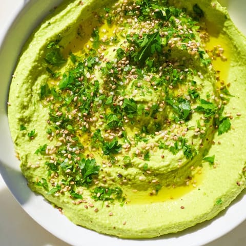 A vibrant bowl of Edamame Hummus with a smooth, pale green texture, garnished with olive oil and sesame seeds, next to colorful vegetable sticks for dipping.  